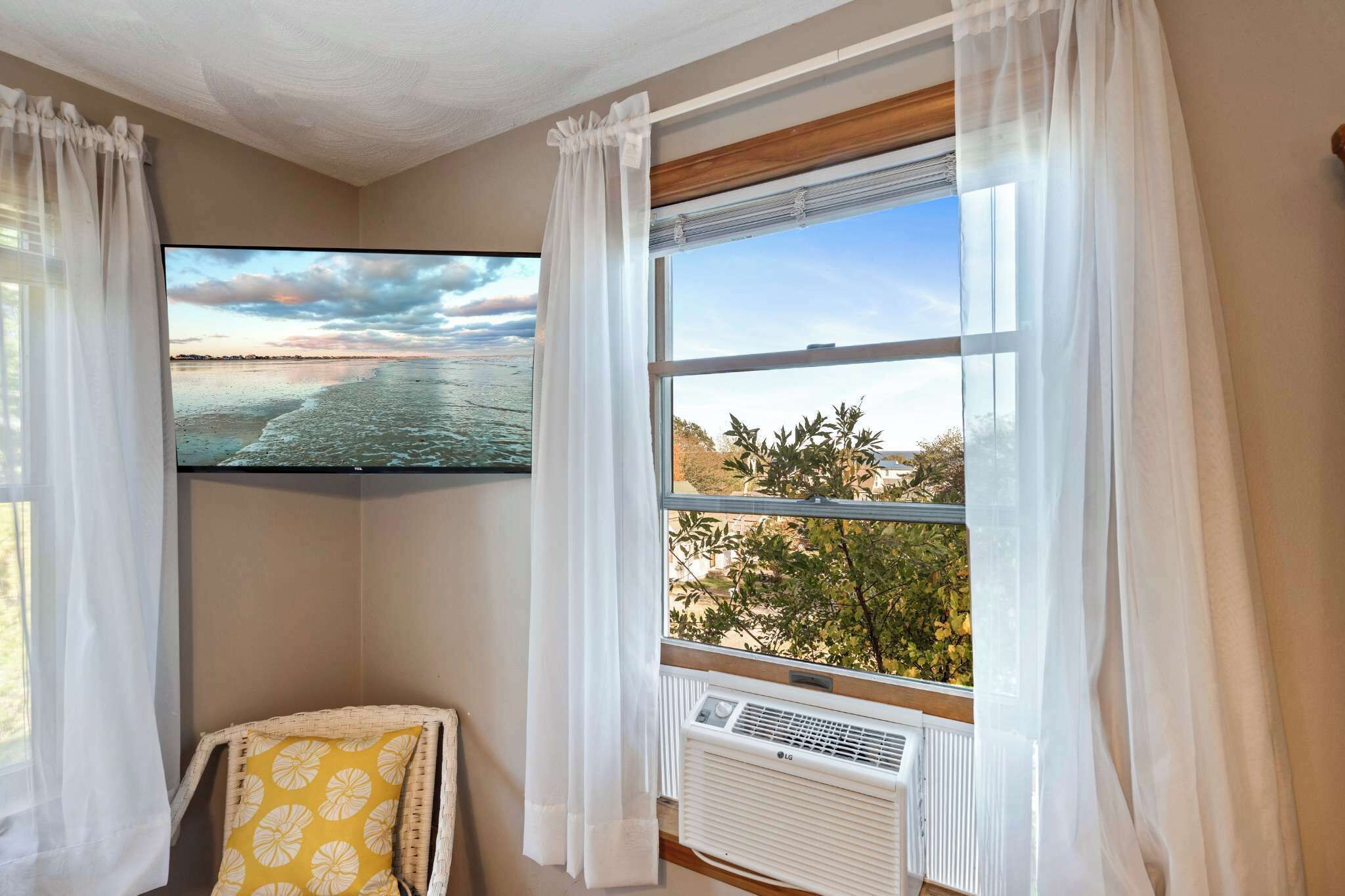 A room with a large window, an air conditioning unit, a wicker chair with a yellow cushion, and a flat-screen TV displaying a beach scene.