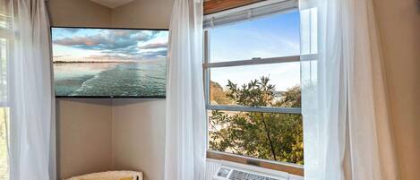 A room with a large window, an air conditioning unit, a wicker chair with a yellow cushion, and a flat-screen TV displaying a beach scene.