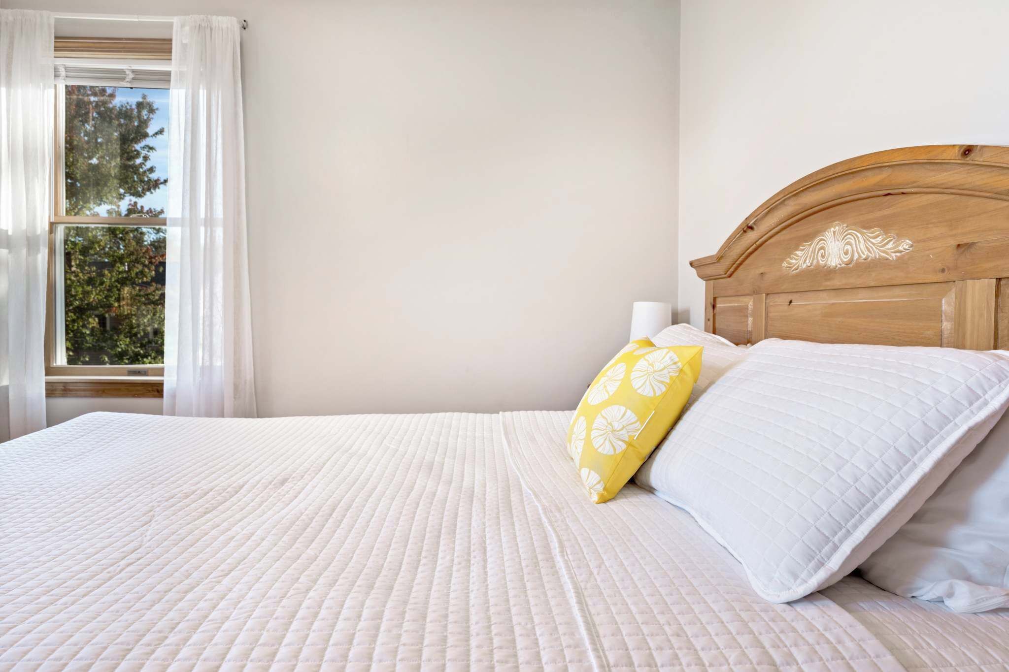 A bed with a quilted white bedspread, a wooden headboard, and two pillows, one yellow with a white pattern and one white.