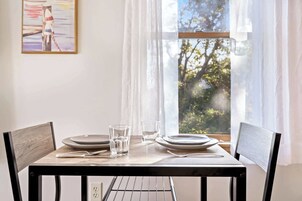 Una mesa de comedor con platos, vasos y cubiertos, frente a una ventana con vistas a árboles.
