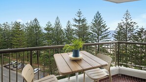 En balkon med et træbord og stole, en potteplante og udsigt til træer og en vej.