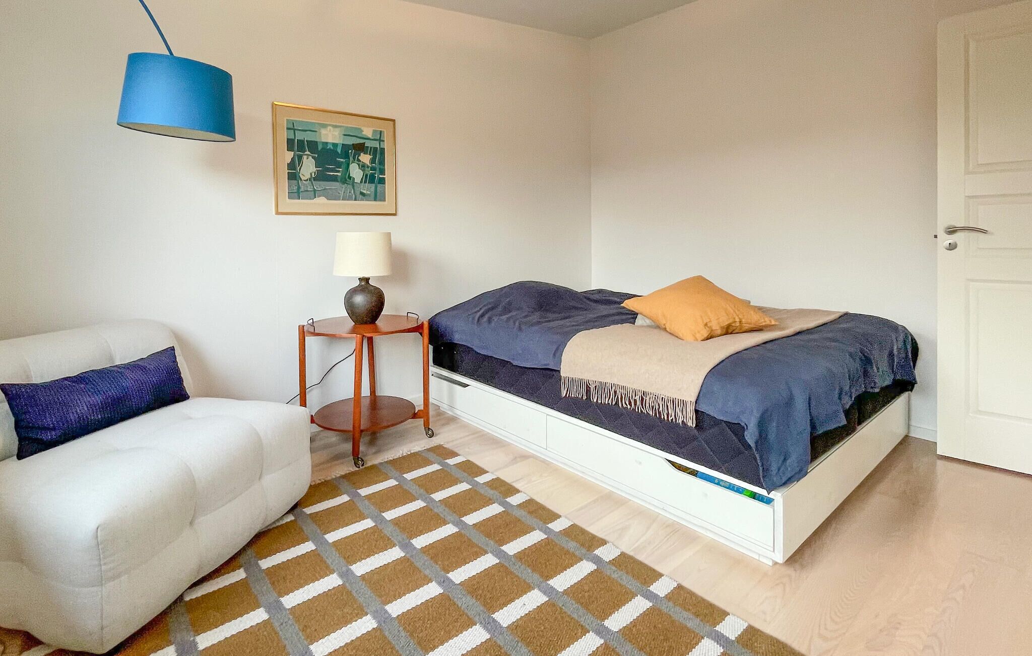 A bedroom with a bed, a chair, a small table with a lamp, a framed picture on the wall, and a patterned rug.