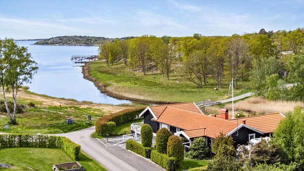 Ett hus vid kusten med ett rött tegeltak, omgivet av frodig grönska och en vattenyta.