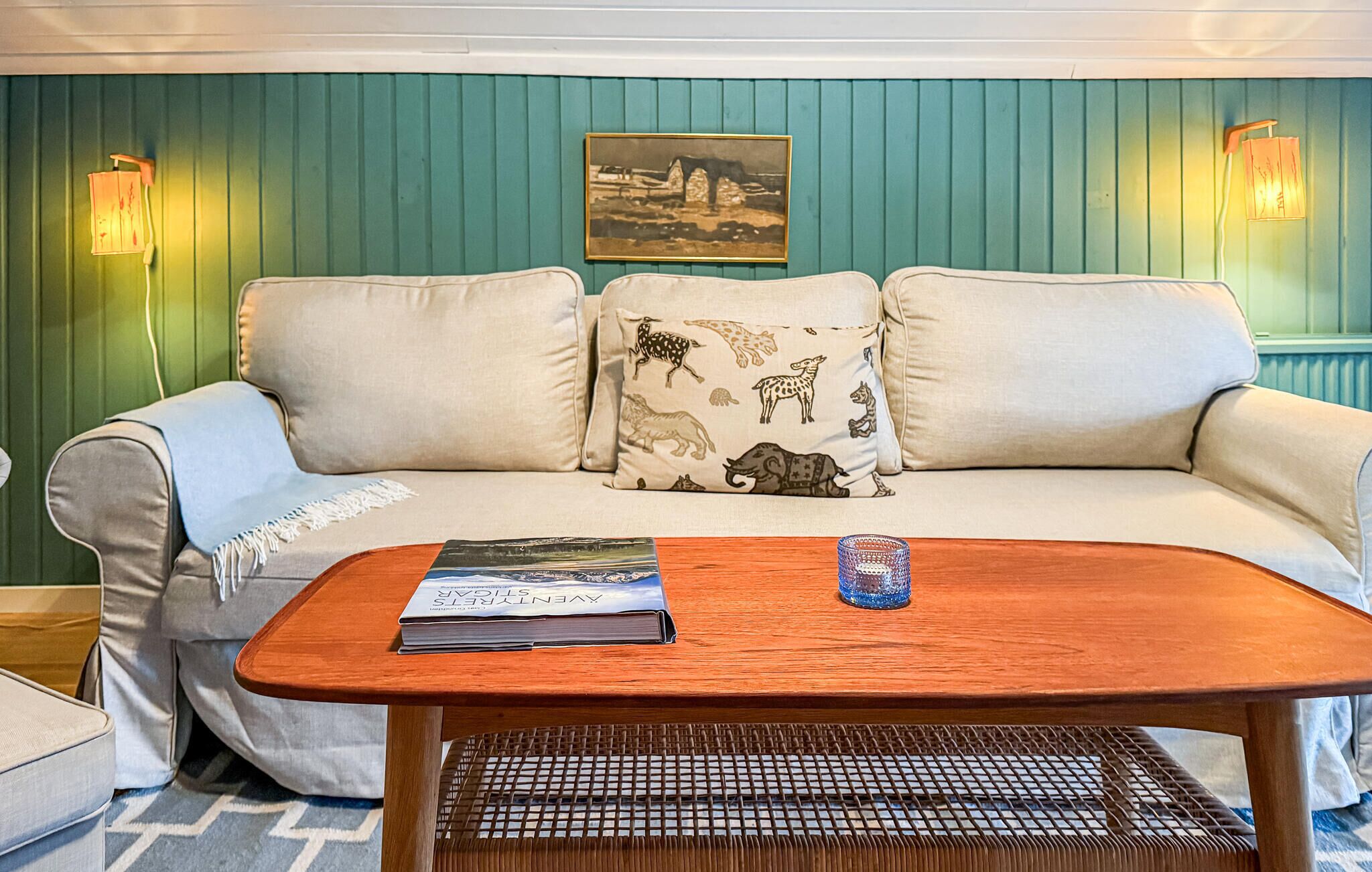 A room with a teal wall, a white sofa with patterned cushions, a wooden coffee table with a magazine, and two wall-mounted lamps.