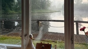 Une vue à travers une fenêtre donnant sur une terrasse en bois surplombant une étendue d’eau, avec un petit nichoir et un objet décoratif posés sur le rebord de la fenêtre.