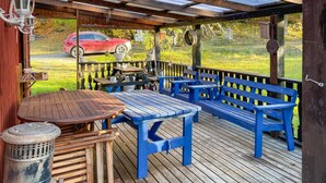 Eine Holzdielen-Terrasse mit blauen Bänken und einem Tisch, umgeben von einem Holzzaun und einem roten Auto im Hintergrund.