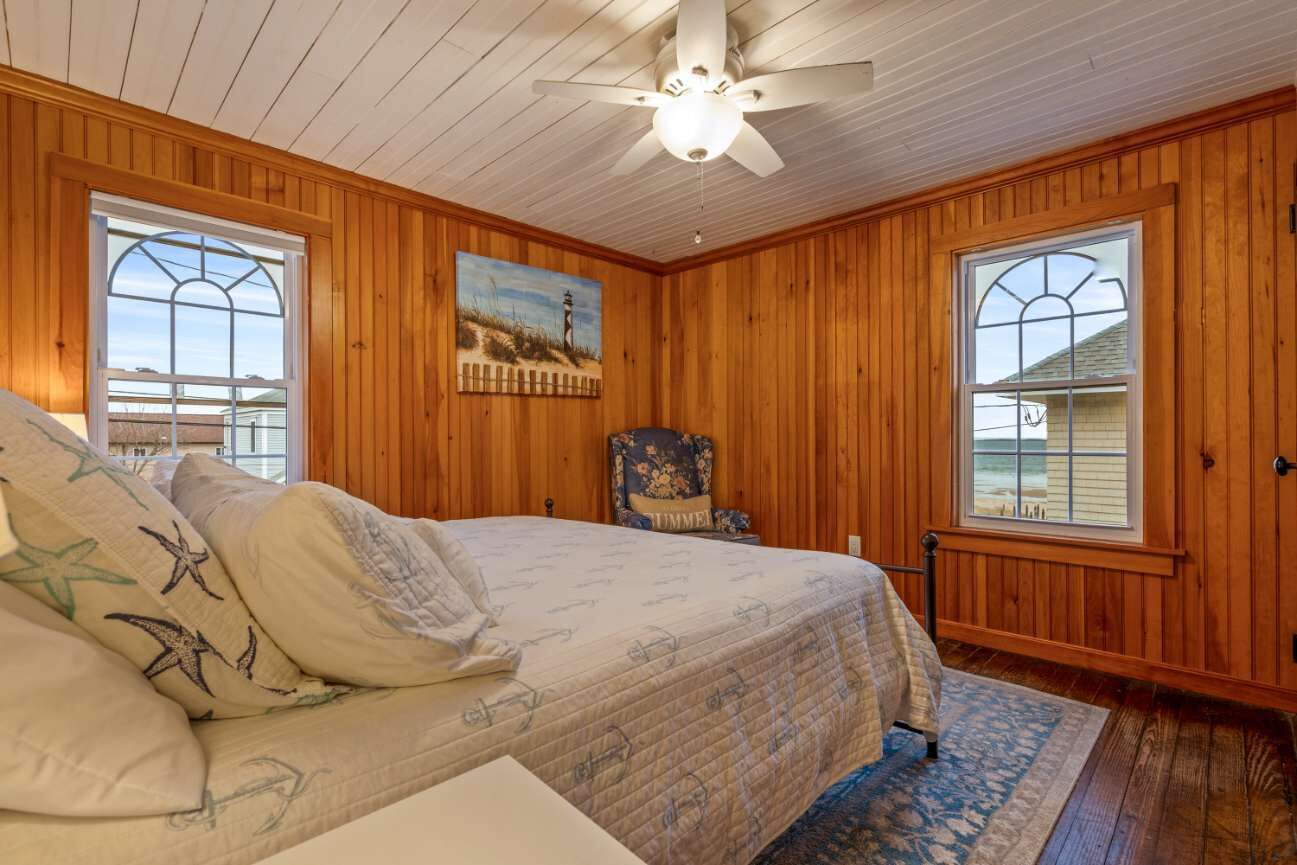 A bedroom with wooden walls, a bed with a quilt, a ceiling fan, and two windows.