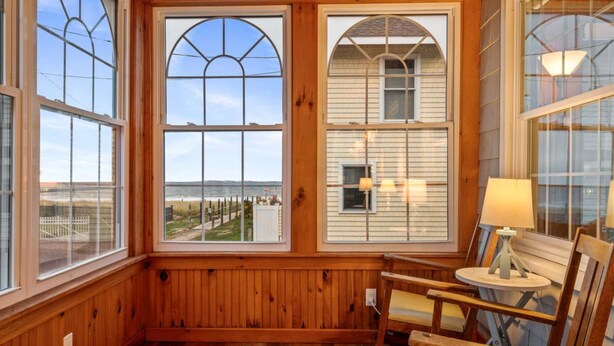 A wooden-paneled room with large arched windows overlooking a beach and a white building. There are two wooden chairs and a small table with a lamp. - Danemead Cottage - For 10ppl (Old Orchard Beach)
