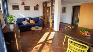 Una sala de estar con un sofá, una mesa de centro y una mesa de comedor con sillas. Hay una planta en maceta sobre el sofá y un frutero en la mesa de comedor.