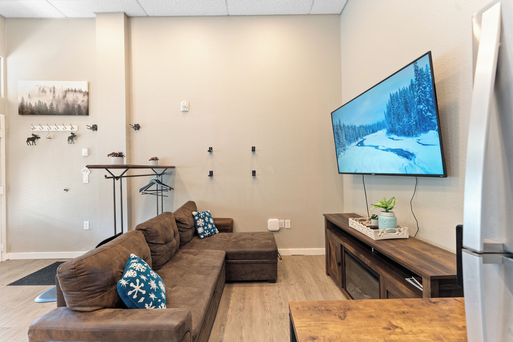 Una sala de estar con un sofá seccional marrón, un televisor de pantalla plana montado en la pared, una mesa de centro de madera y un mueble pequeño.