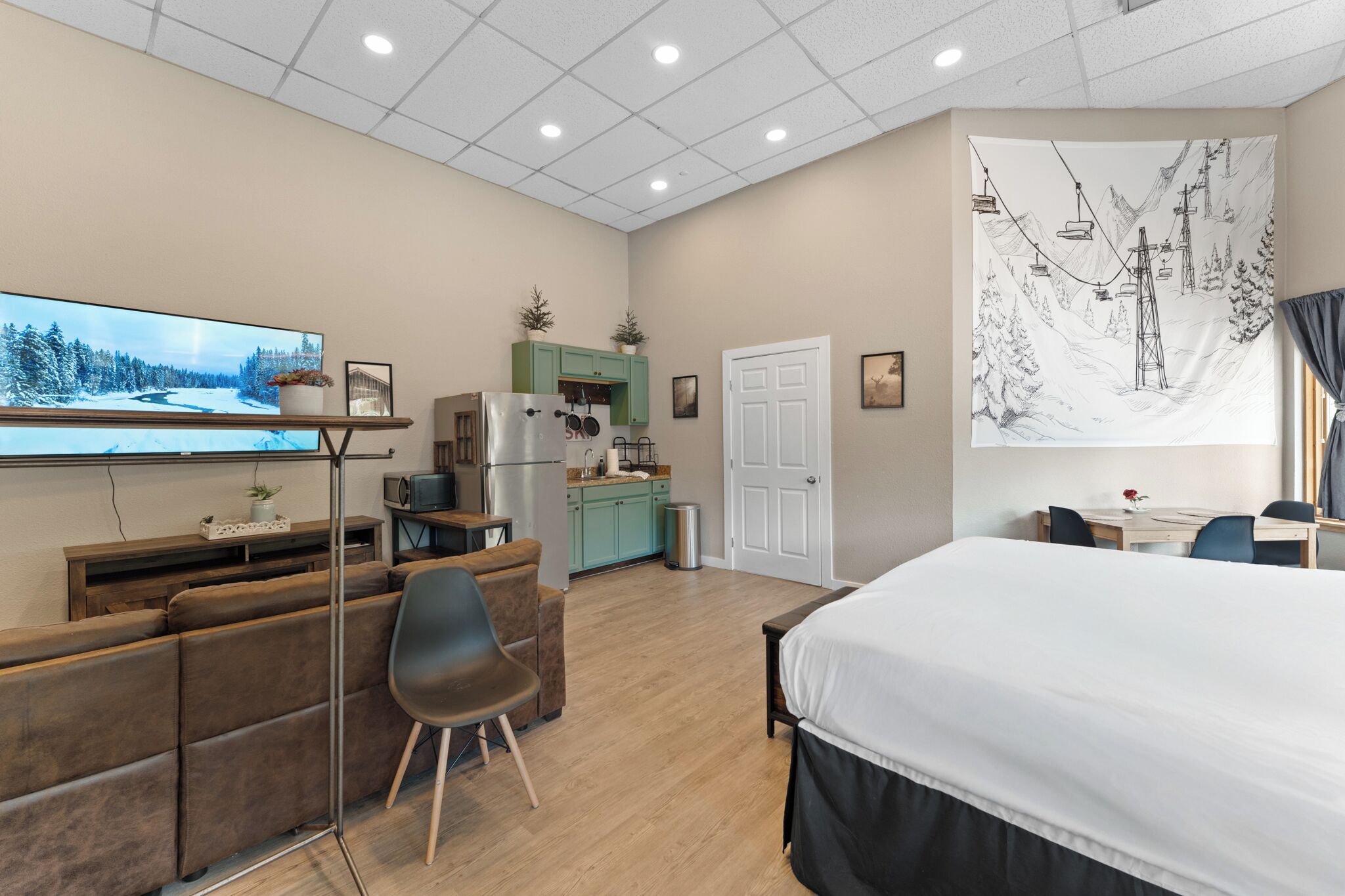 Una habitación de hotel moderna con una cama grande, televisión, kitchenette y una zona de estar con sofá y sillones.