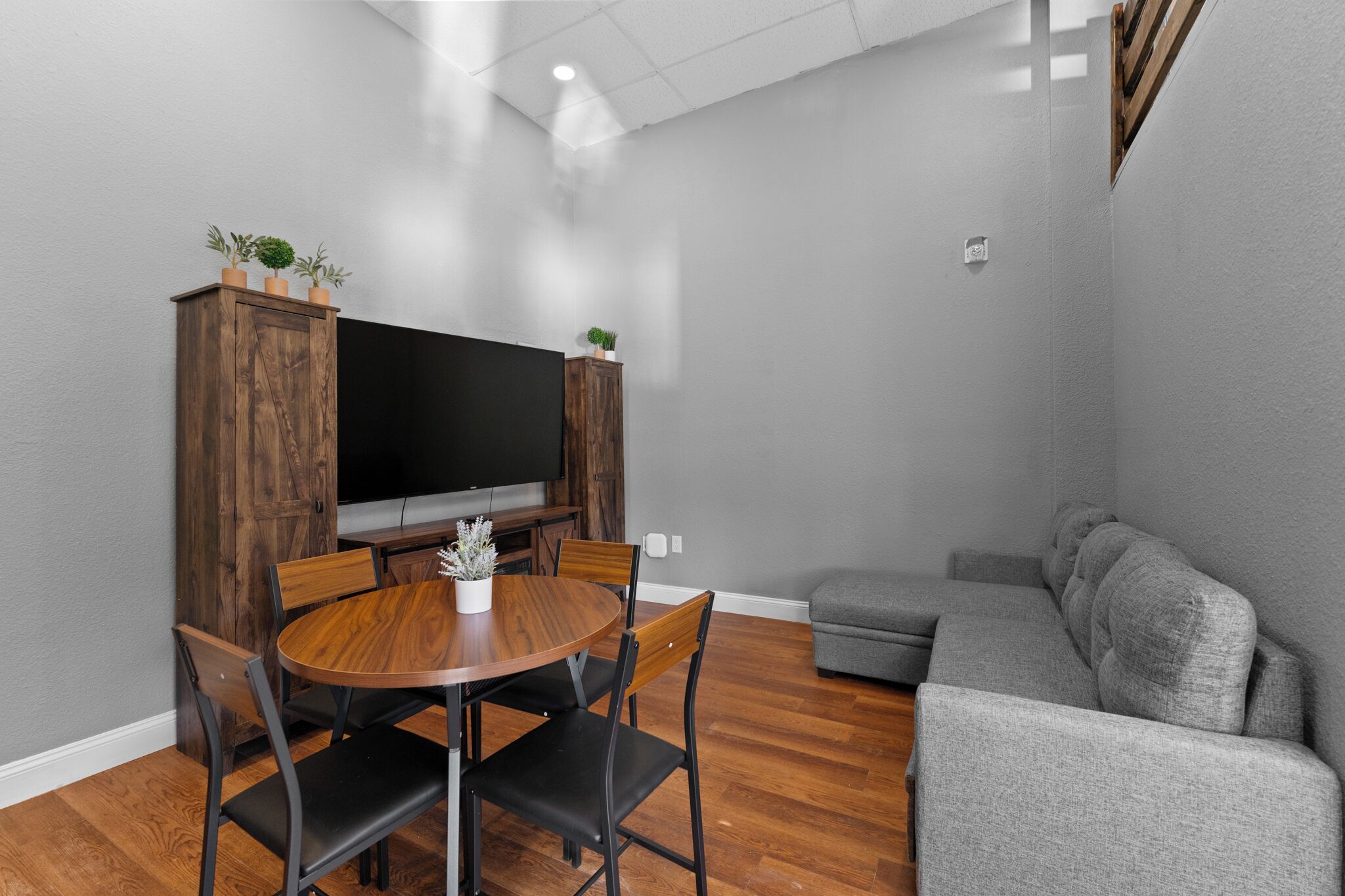 A modern living room with a wooden dining table, grey sofa, and a flat-screen TV on a wooden cabinet.