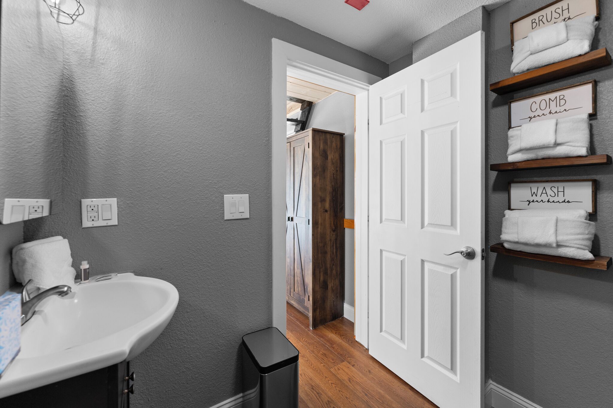 A bathroom with a white sink, a trash bin, and shelves with towels labeled "BRUSH," "COMB," and "WASH."