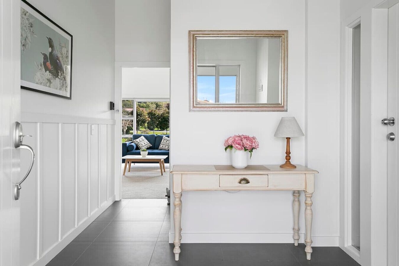 Een gang met een witte console tafel, een spiegel, een lamp en een lijst met een afbeelding aan de muur.