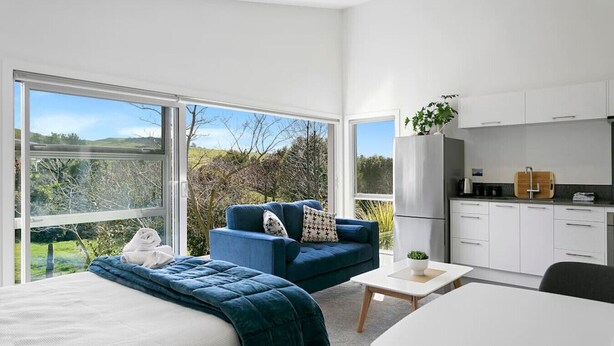 A modern bedroom with a large bed, a blue sofa, and a small table. There is a kitchen area with white cabinets and a countertop.