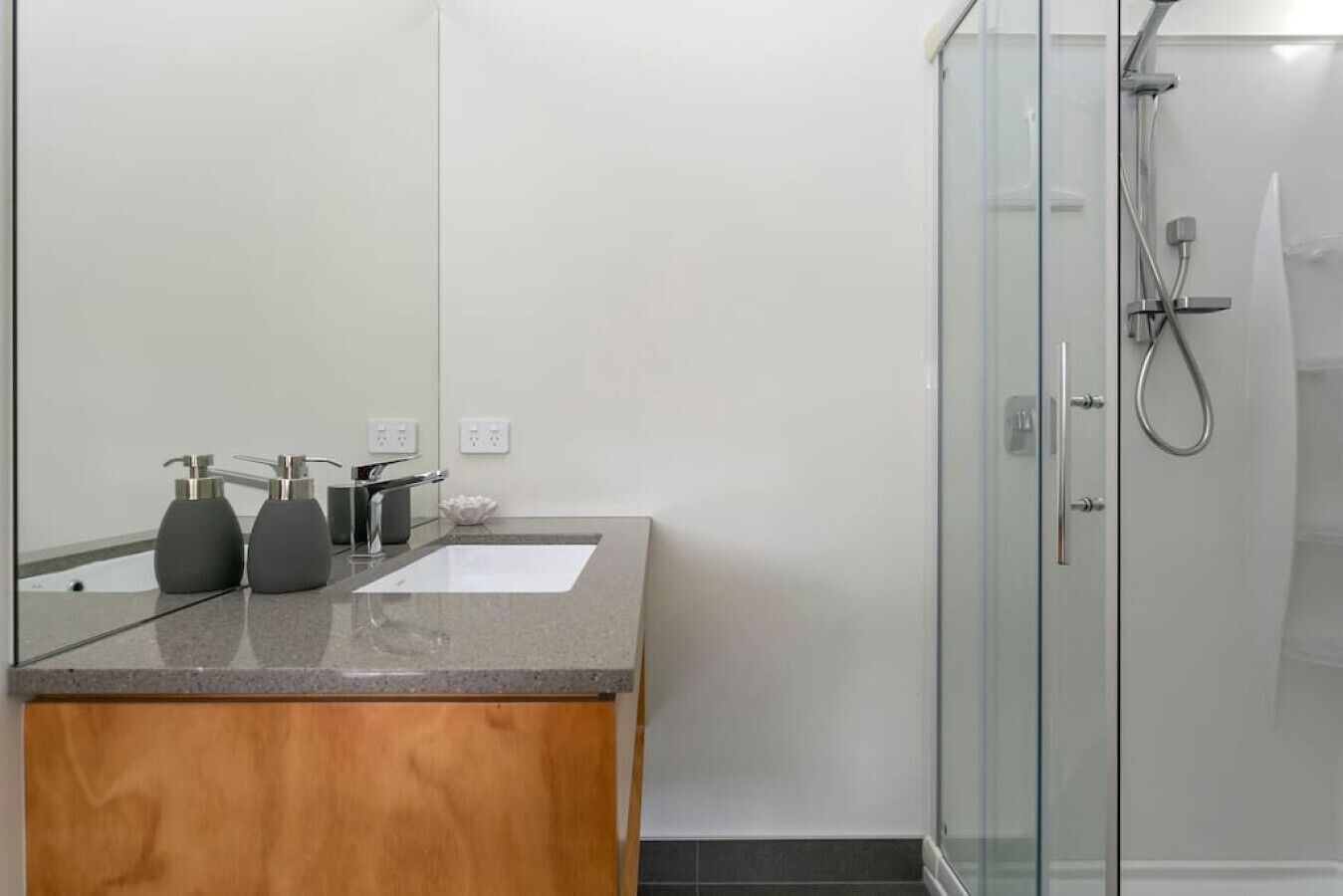 Ein modernes Badezimmer mit einem Holzkosmetikschrank, zwei schwarzen Seifenspendern, einem weißen Waschbecken und einer Dusche mit Glastrennwand.