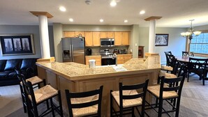 A modern kitchen with granite countertops, wooden cabinets, and stainless steel appliances. There are six bar stools and a dining table with chairs. - Greenspring towhhome- Free shuttle and clubhouse (Dover)