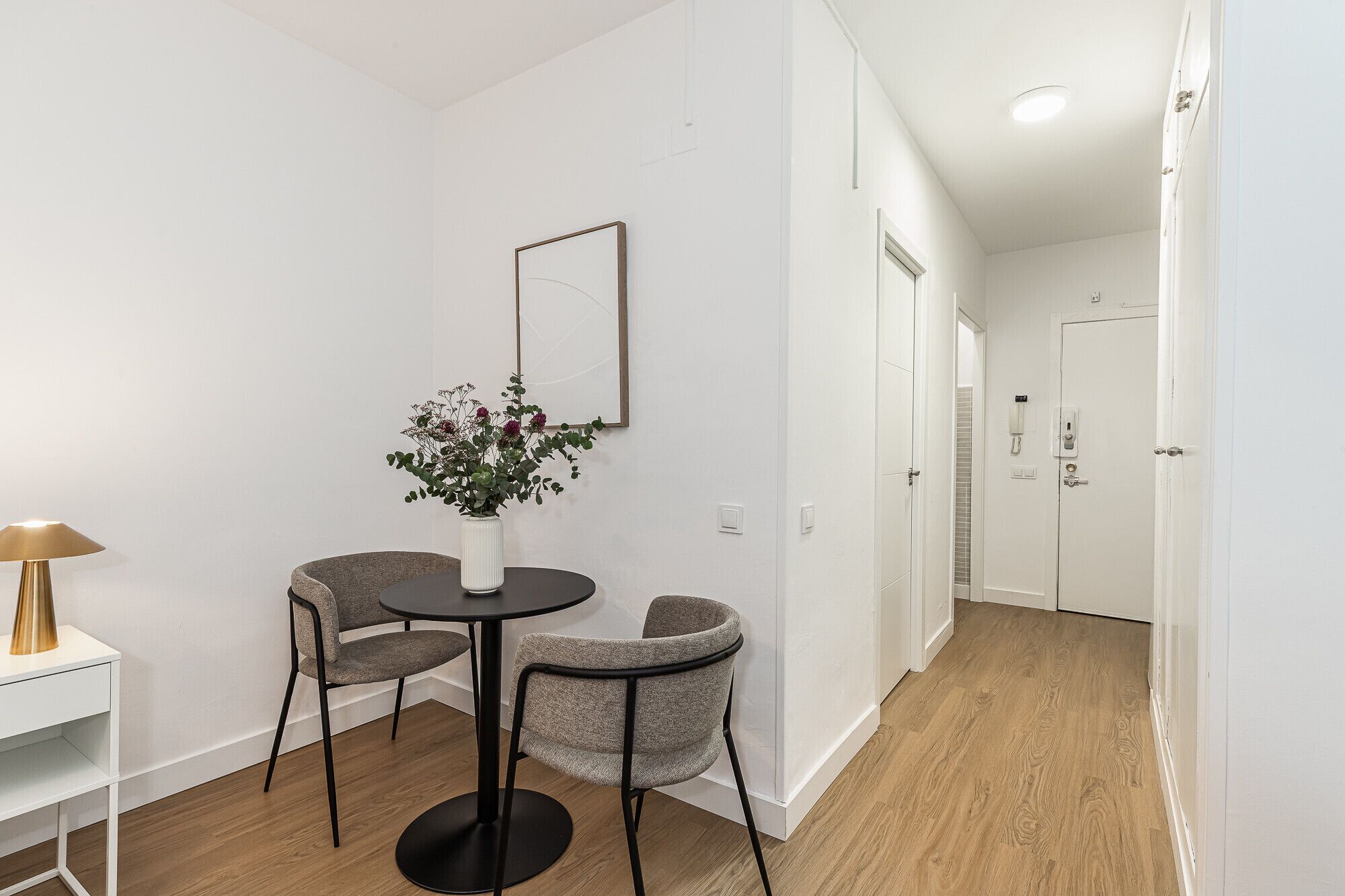 Een moderne, minimalistisch ingerichte kamer met een ronde tafel en twee stoelen, een klein wit tafeltje met een lamp en een vaas met bloemen.