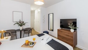 Moderni hotellihuone, jossa on sänky, televisio, ruokailualue pöytineen ja tuoleineen sekä kylpyhuone, joka on näkyvissä avonaisen oven läpi.