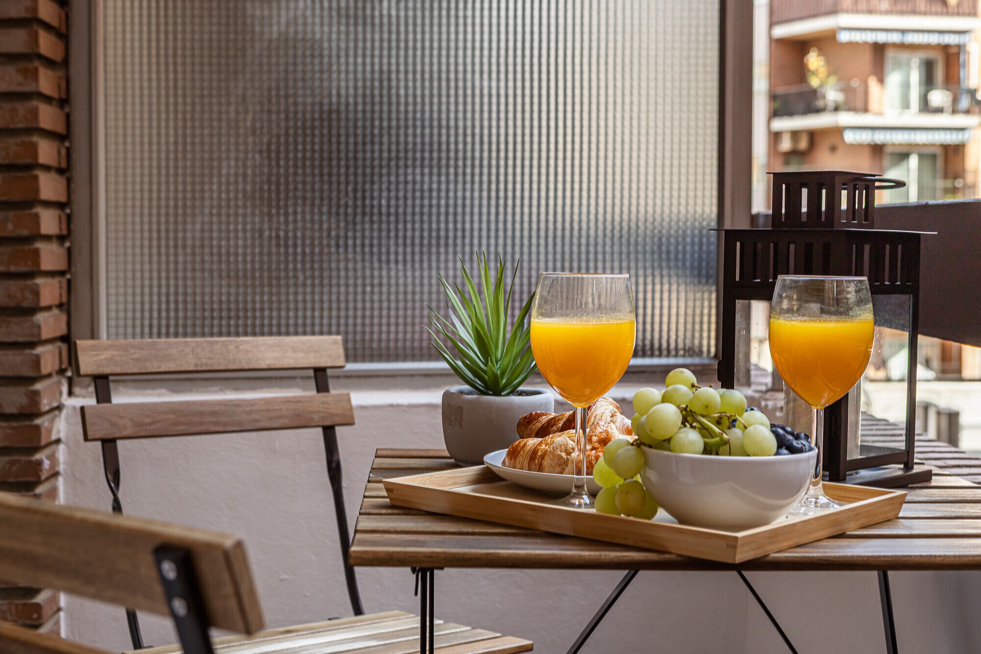 A breakfast tray with croissants, grapes, and a bowl of fruit on a wooden table by a window.