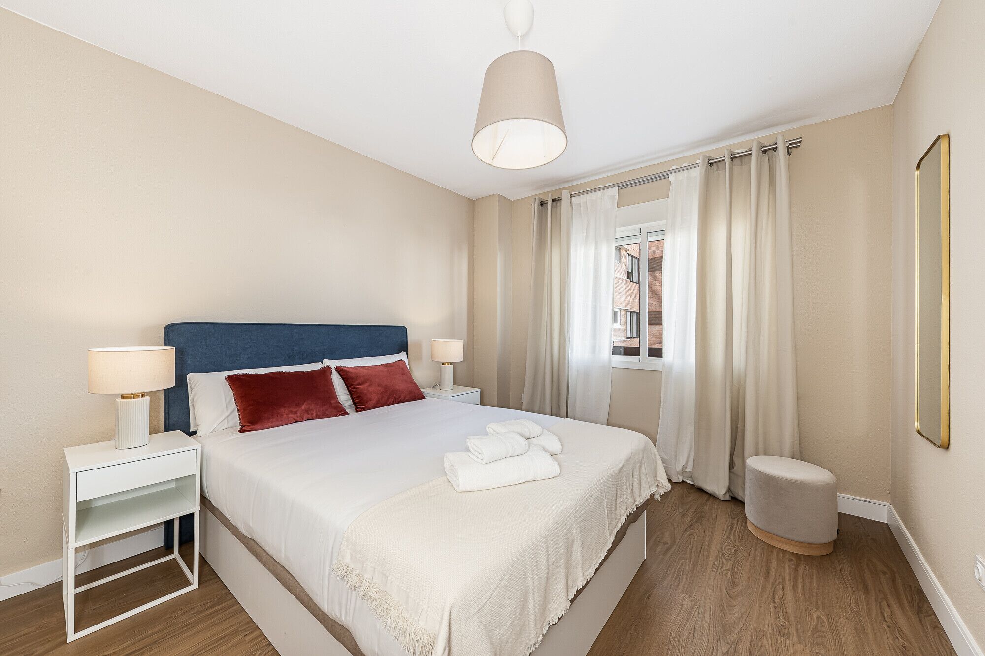 Ein Hotelzimmer mit einem Bett, zwei Nachttischen, einer Hängelampe, einem Fenster mit Vorhängen und einem kleinen Hocker.
