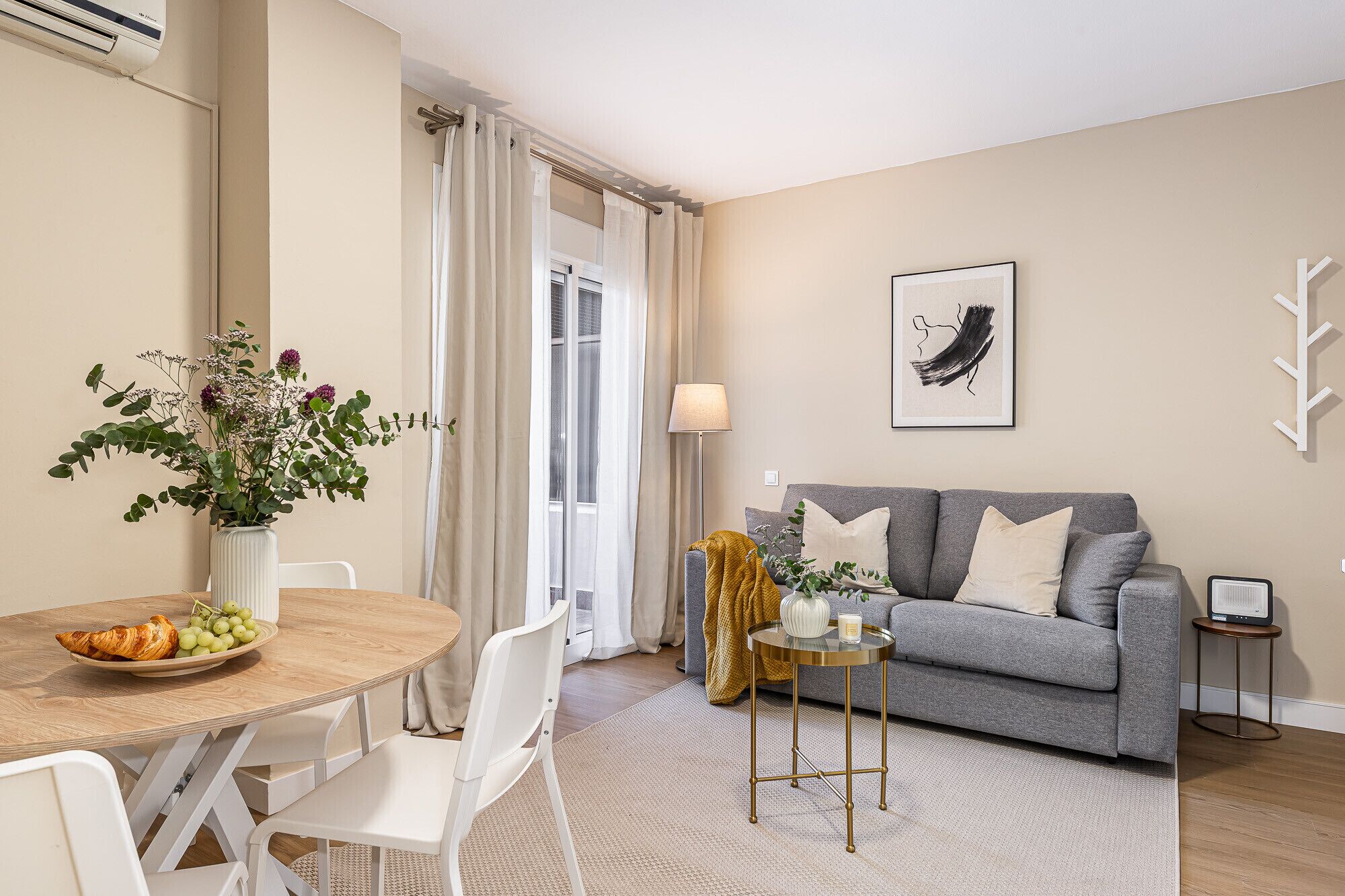 Ein modernes Wohnzimmer mit einem grauen Sofa, weißen Sesseln, einem runden Holztisch mit Blumenvase und einem kleinen Beistelltisch mit Lampe.