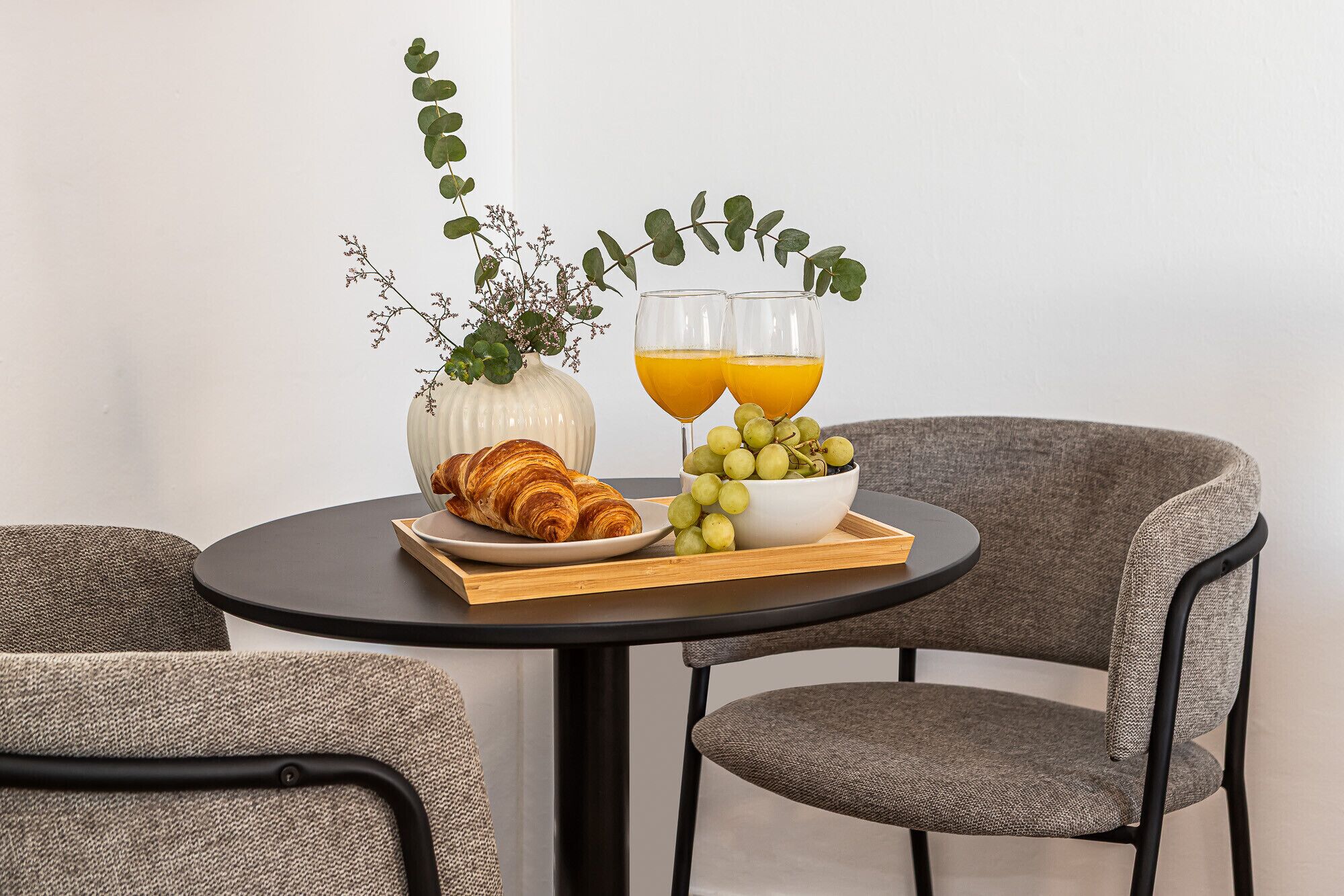 A dining table with a croissant, a bowl of grapes, and two glasses of orange juice, set against a white wall with a plant.