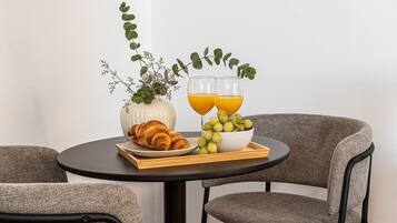 A dining table with a croissant, a bowl of grapes, and two glasses of orange juice, set against a white wall with a plant.