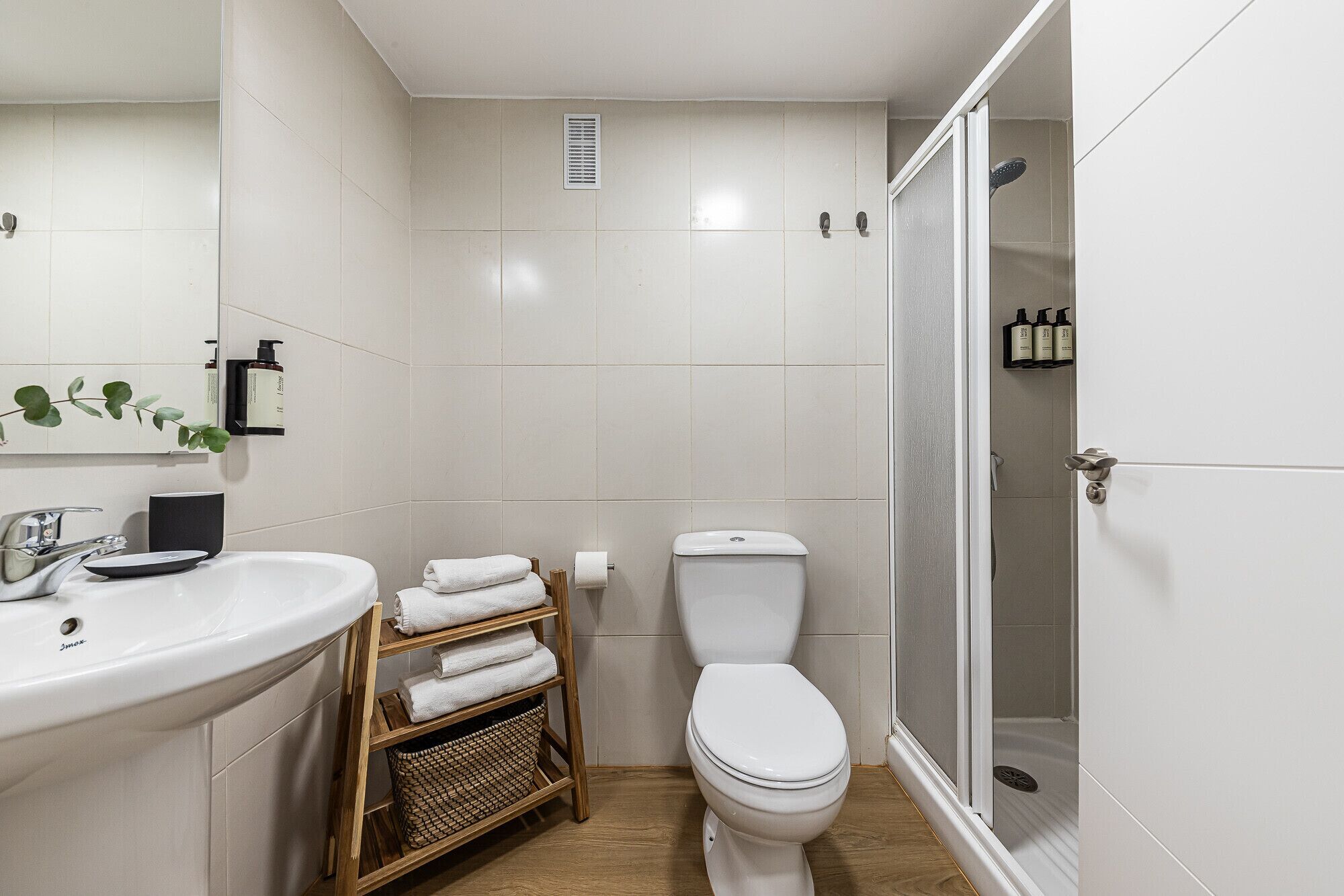 A modern bathroom with a white sink, toilet, and shower. There is a towel rack and a basket on the floor.