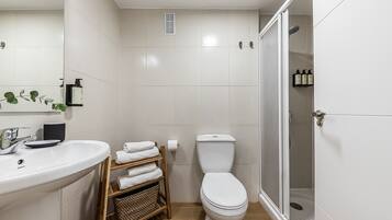 A modern bathroom with a white sink, toilet, and shower. There is a towel rack and a basket on the floor.