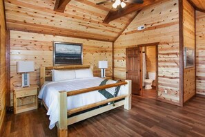 Ein Schlafzimmer einer Holzblockhütte mit einem Bett, Nachttischen, einem Deckenventilator und Blick ins Freie.