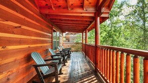 A wooden deck with a railing, benches, and a view of the surrounding forest. - Outdoor Theater + Jetted Hot Tub | 8BR Sleeps 24 (Sevierville)
