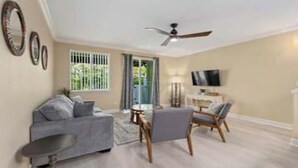 Una sala de estar con sofá, sillón y mesa de centro. Hay un televisor y un ventilador de techo.