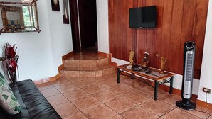 A living room with a black leather sofa, a coffee table with decorative items, a TV mounted on a wooden wall, and a standing heater. - Family room 2 (Manuel Antonio)