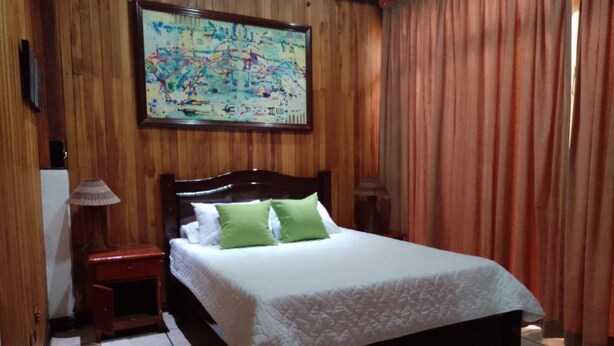 A bedroom with a wooden headboard, a bed with white bedding and green pillows, a nightstand with a lamp, and a painting on the wall.