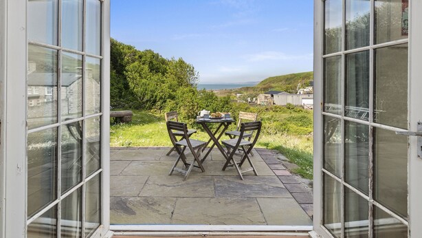 Puerta abierta que da a un patio con mesa y sillas, con vistas a un paisaje pintoresco con colinas y una masa de agua.