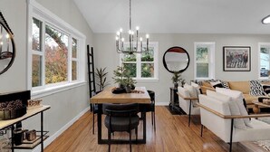A dining area with a wooden table, black chairs, and a chandelier. There are windows with autumn foliage visible outside.