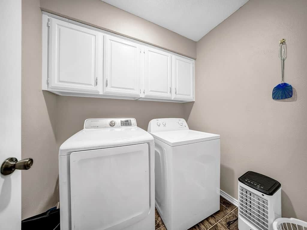 A laundry room with two white front-loading washers and dryers, a wall-mounted fan, and a broom hanging on the wall.