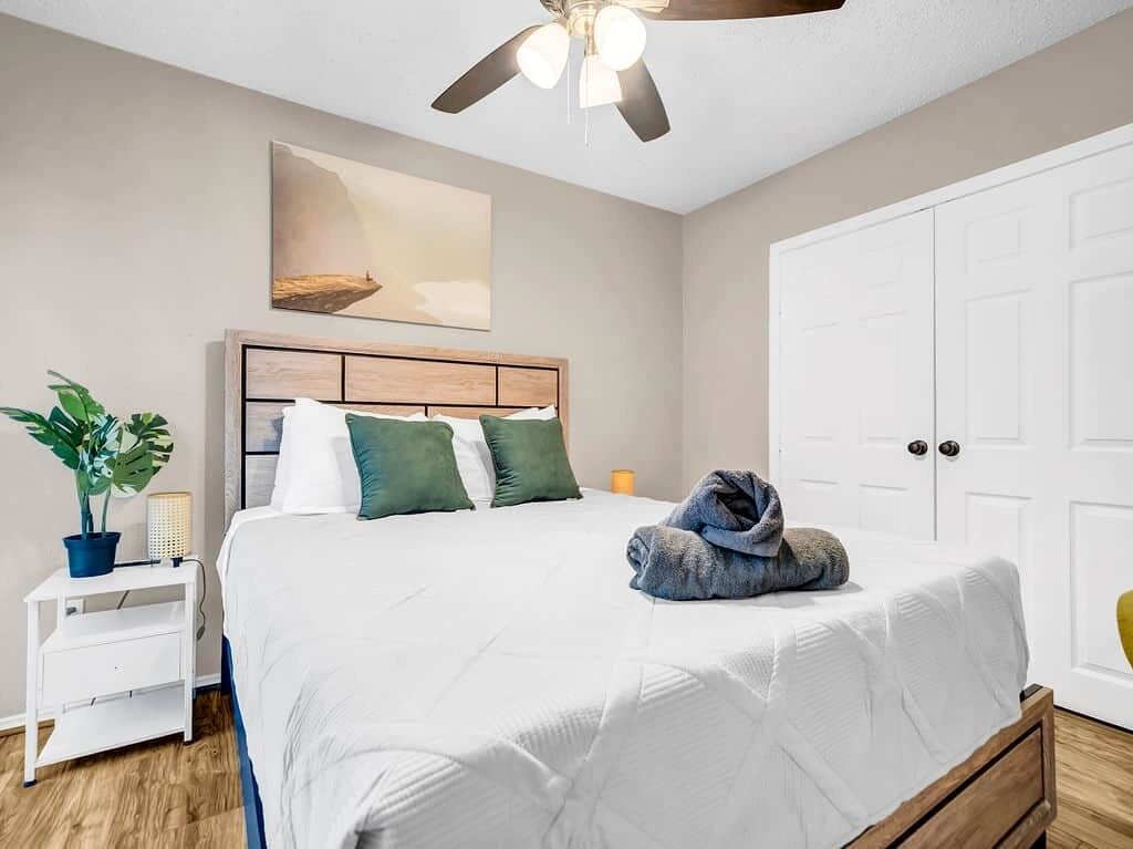 Un dormitorio con cama, ventilador de techo, mesita de noche con planta y una puerta.