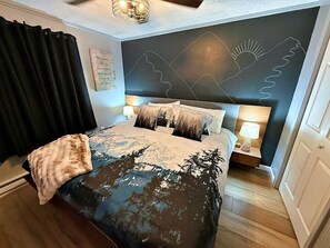 Une chambre à coucher avec un lit, une table de chevet, un mur décoré d’un motif de montagne et une fenêtre avec des rideaux.