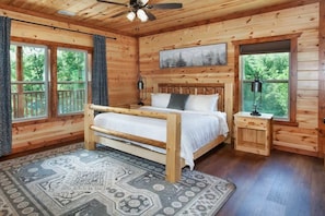 Ein Schlafzimmer in einer Holzhütte mit einem Bett, einem Nachttisch, einem Deckenventilator und großen Fenstern.
