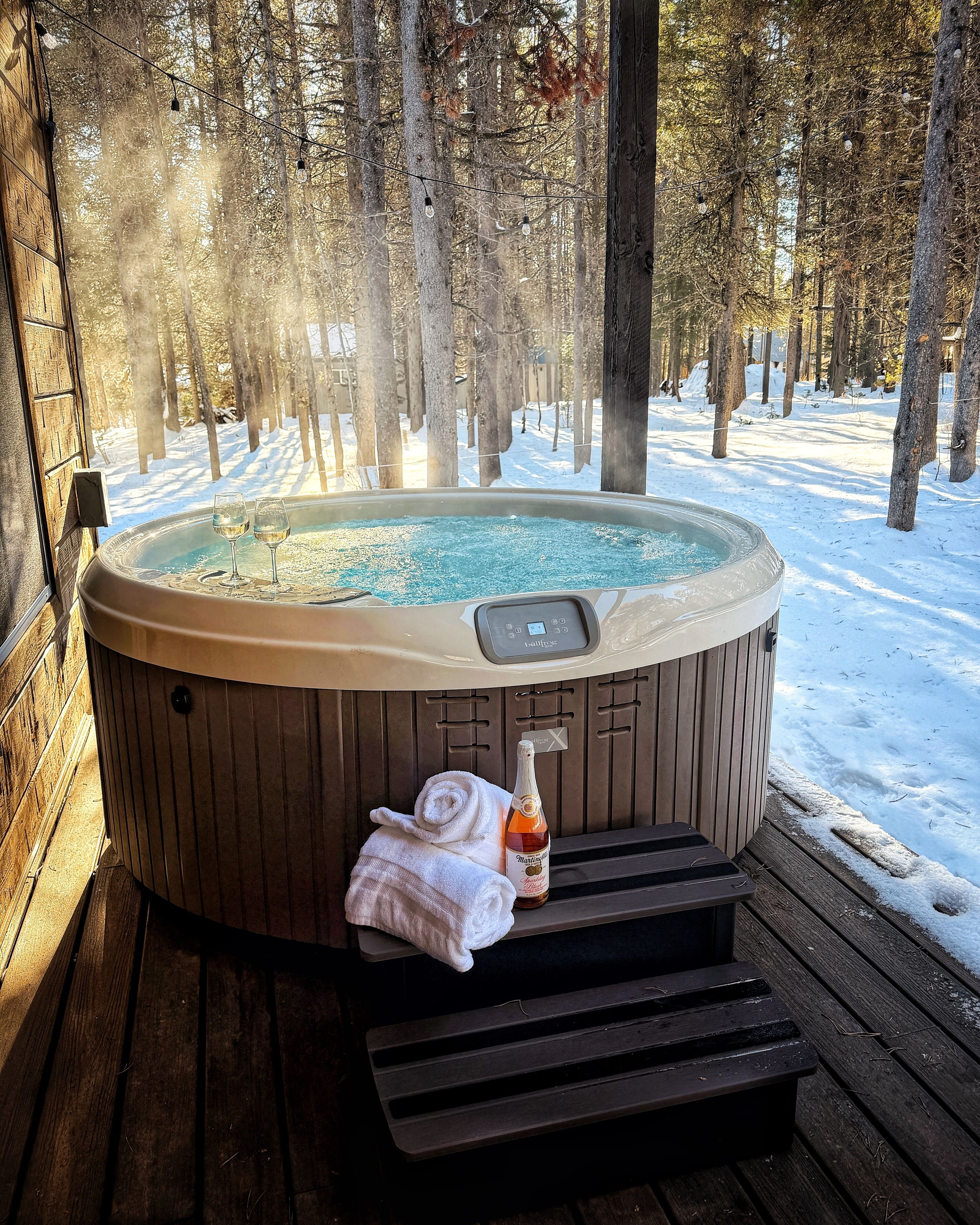 A hot tub with a control panel, a bottle of champagne, and a towel on a wooden deck surrounded by snow and trees.