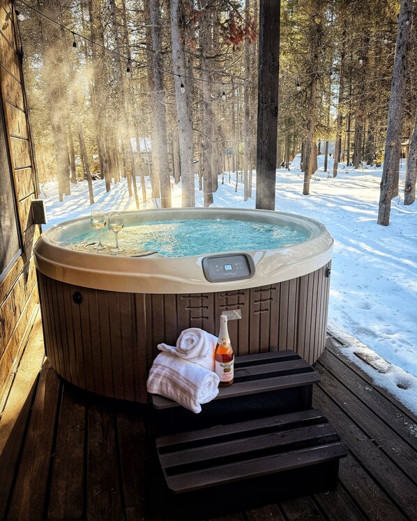 Un jacuzzi con panel de control, una botella de champán y una toalla sobre una terraza de madera rodeada de nieve y árboles.