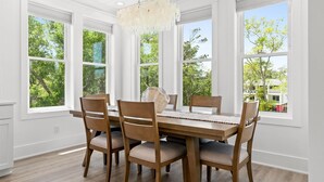 Comedor con mesa y sillas de madera, ventanas amplias y una araña.