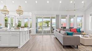 Una cocina amplia con una isla grande, un comedor con un sofá gris y cojines coloridos, y un balcón con vistas al vecindario.