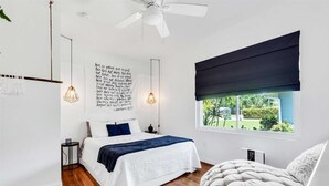 A bedroom with a bed, bedside tables, a ceiling fan, a window with a view, and a wall with text. - Tangerine House | Fire pit + Tree House (Sarasota)