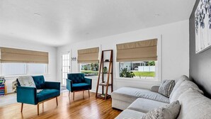 A spacious living room with a wooden floor, a teal armchair, a grey sofa, and a tall floor lamp. - Tangerine House | Fire pit + Tree House (Sarasota)