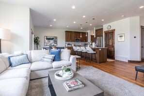 A modern living room with a sectional sofa, a coffee table with magazines, and a kitchen area with wooden cabinets and a bar stool. - Summer by the Lake! Beautiful Swimming Pool, Patio w Hot Tub and BBQ Nature Trails Hiking Biking (Hideout)