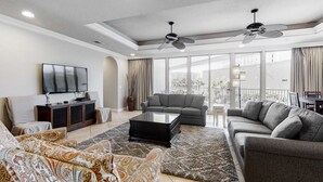 A spacious living room with a grey sofa, a patterned armchair, a coffee table, and a flat-screen TV on a wooden cabinet. There are two ceiling fans and large windows with curtains. - Stylish Gulf-Front Condo w/ Balcony & Pools (Miramar Beach)