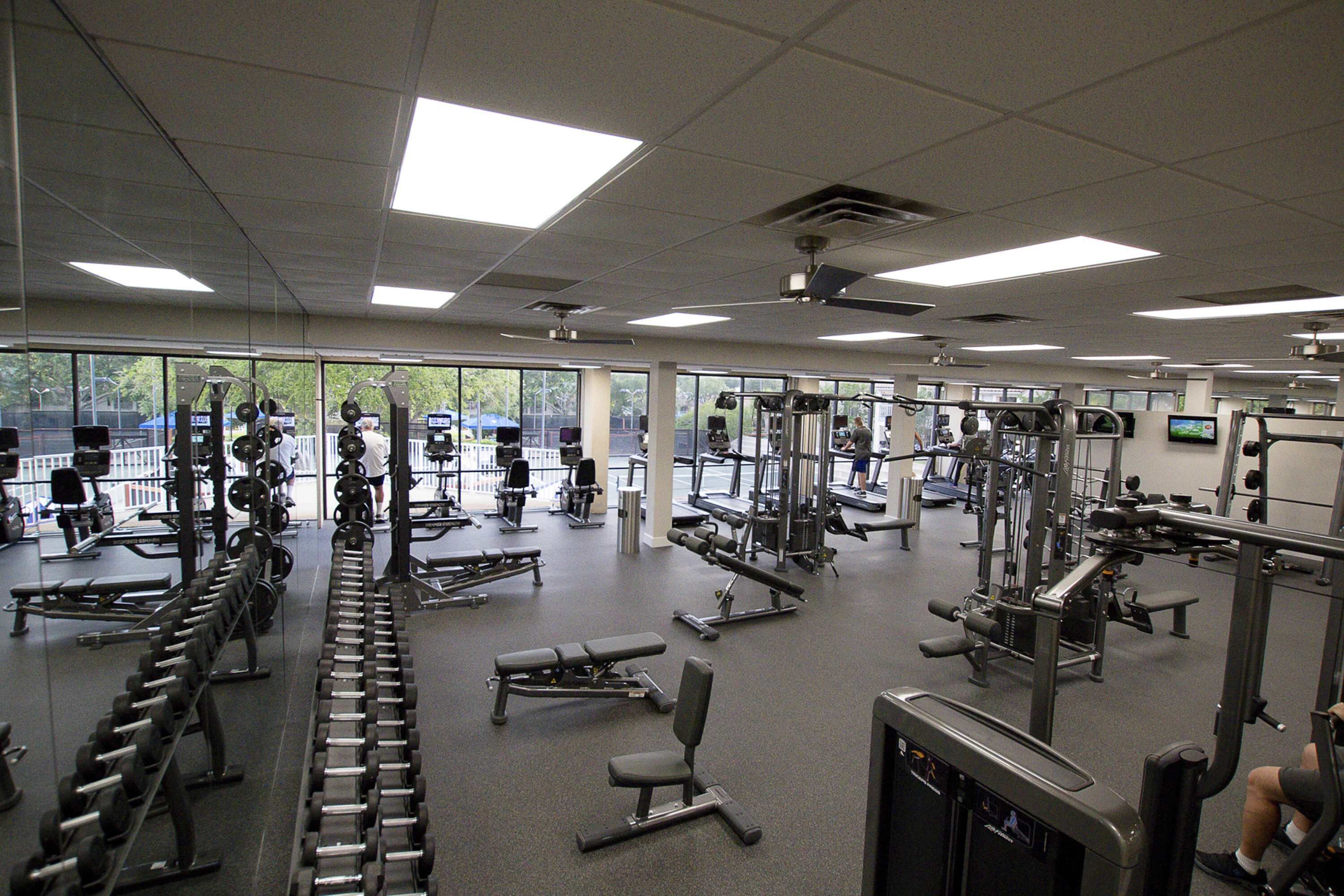 Een sportschool met diverse trainingsapparaten, grote ramen en een plafondventilator.